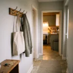 a front hallway at dusk, a bag hanging on a hook, keys on a small shelf, a warm light at the far end where the kitchen is; no people. Alt: Front hallway in the early evening with a bag on a hook and keys on a shelf.